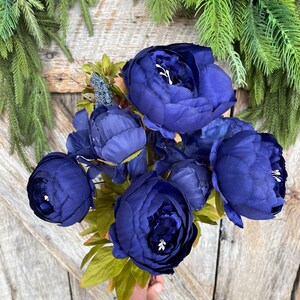 May include: A bouquet of deep blue artificial peonies with green leaves. The flowers are clustered together, held by a hand, against a rustic wooden background. Greenery frames the top of the image.