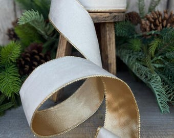May include: A roll of ivory-colored ribbon with a gold metallic trim. The ribbon is displayed on a small wooden stool, with a portion unrolled to show the gold underside. The background includes pine branches and cones, suggesting a holiday theme.