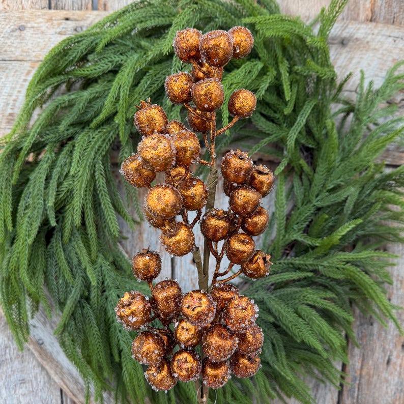 May include: A decorative floral stem featuring numerous round, metallic copper-colored berries with a textured, glittery finish. The berries are clustered along a central brown branch, creating a dense, spherical arrangement of small, shiny, spherical elements.
