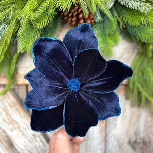 May include: A large, navy blue velvet flower with glitter edging and a textured center. The flower is held in front of a backdrop of evergreen branches and a pinecone. The flower's petals are layered, creating a full, dimensional look.