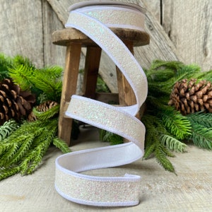 May include: A roll of white ribbon with a glittery, iridescent surface and a white satin backing. The ribbon is displayed on a small wooden stool, with evergreen branches and pine cones in the background. Perfect for holiday crafts and gift wrapping.
