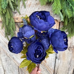 May include: A bouquet of deep blue artificial peonies with green leaves. The flowers are in full bloom, with multiple petals visible. The bouquet is held against a wooden background, with green foliage in the background.