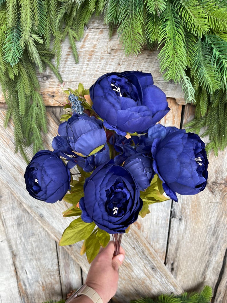 May include: A bouquet of deep blue artificial peonies with green leaves. The flowers are in full bloom, with multiple petals visible. The bouquet is held against a wooden background, with green foliage in the background.