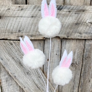 May include: Three white fluffy pom-pom bunny decorations with pink ears. The bunny decorations are on white stems.