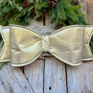 May include: A large gold bow made of fabric with a satin finish. The bow is displayed on a wooden surface.