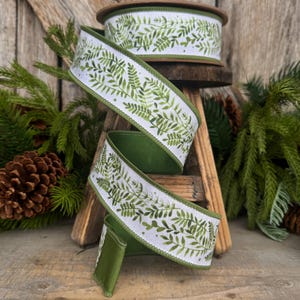 May include: A spool of white ribbon featuring a green watercolor fern leaf pattern. The ribbon has a solid green stitched edge and is displayed in a cascading spiral. The design consists of various shades of green foliage printed on a white fabric base.