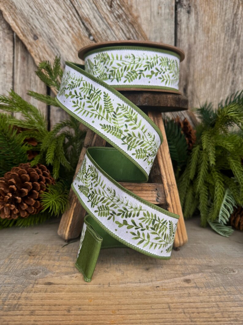 May include: A spool of white ribbon featuring a green watercolor fern leaf pattern. The ribbon has a solid green stitched edge and is displayed in a cascading spiral. The design consists of various shades of green foliage printed on a white fabric base.