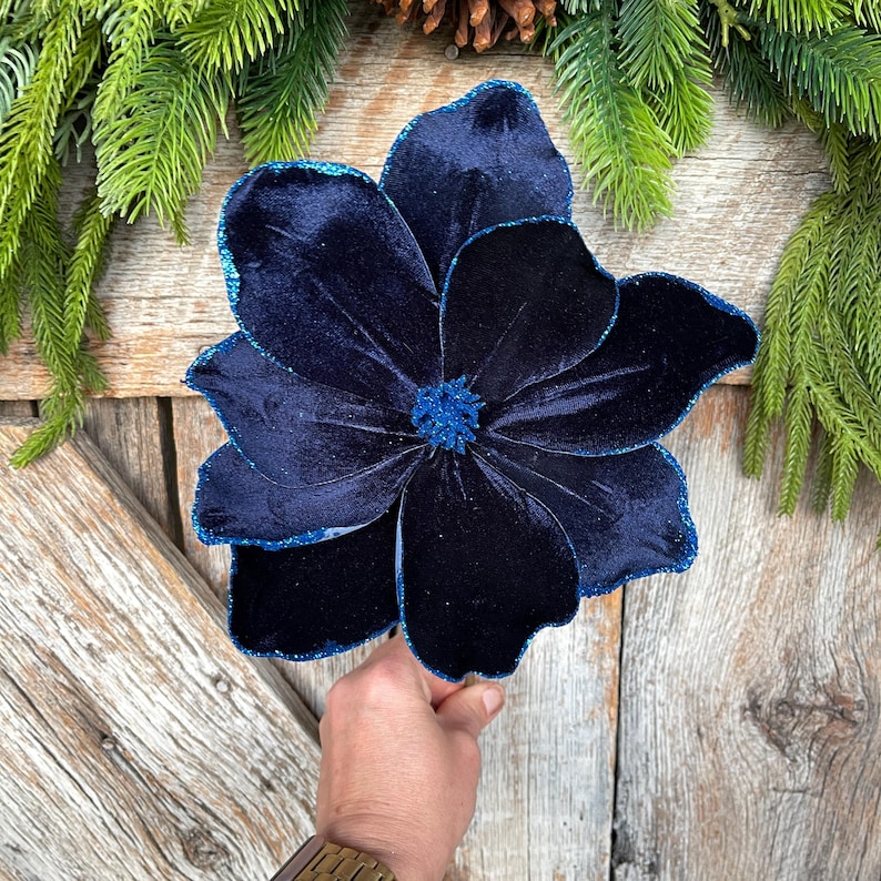May include: A large, navy blue velvet flower with glitter trim. The flower has multiple petals and a textured center. The flower is held against a wooden background with pine needles and a pine cone.