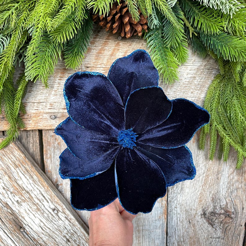 May include: A large, navy blue velvet flower with glittered edges. The flower has multiple petals and a textured center. The background includes evergreen branches and a pinecone, suggesting a holiday or seasonal theme.