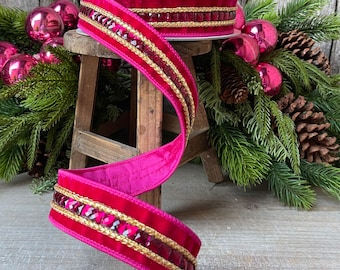 Puede incluir: Un rollo de cinta de terciopelo magenta vibrante con un borde decorativo dorado y joya. La cinta se muestra sobre un pequeño taburete de madera, con adornos navideños y ramas de pino en el fondo. Ideal para manualidades y envolver regalos.