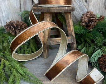 May include: A spool of brown velvet ribbon with a gold trim sits on a small wooden stool. The ribbon is unrolled, revealing a gold underside. Pine needles and pine cones surround the ribbon, suggesting a holiday theme.