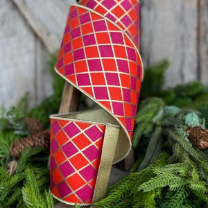 May include: A roll of decorative ribbon with a vibrant geometric pattern. The ribbon features a repeating diamond pattern in shades of red and fuchsia, accented with gold lines. The edges are trimmed with gold. The ribbon is displayed on a wooden stand with greenery.