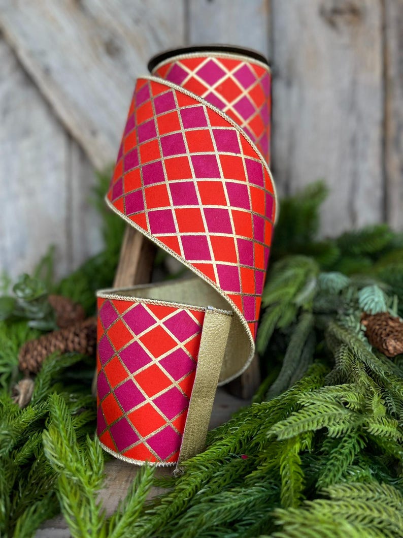 May include: A roll of decorative ribbon with a vibrant geometric pattern. The ribbon features a repeating diamond pattern in shades of red and fuchsia, accented with gold lines. The edges are trimmed with gold. The ribbon is displayed on a wooden stand with greenery.