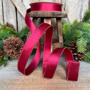 May include: A spool of deep red satin ribbon sits atop a small wooden stool, with a length of ribbon cascading down. The ribbon has a subtle sheen and a stitched edge. Pinecones and greenery are in the background.