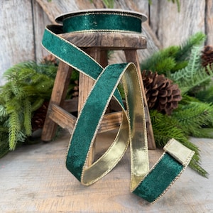 May include: A spool of dark green velvet ribbon featuring a metallic gold edge. The ribbon is wrapped around a circular core and cascades downward, displaying a soft, textured green surface on one side and a smooth, reflective gold finish on the other.