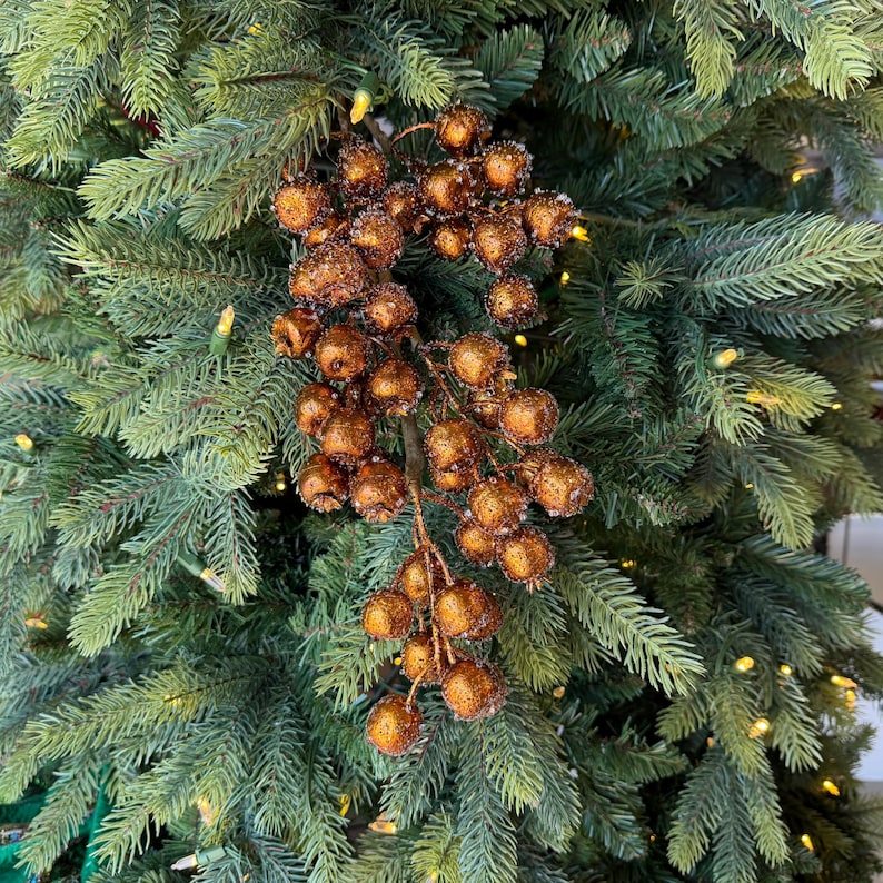 May include: A decorative branch featuring numerous round, copper-colored berries with a textured, glittery finish. The cluster of berries is attached to a central stem and is displayed against a background of green artificial pine tree needles.