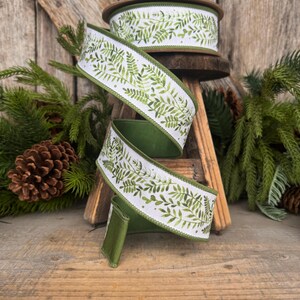 May include: A spool of wired ribbon featuring a white background with a repeating green watercolor fern leaf pattern. The ribbon has solid green finished edges and is coiled in a spiral shape, showcasing the botanical print on the front side.