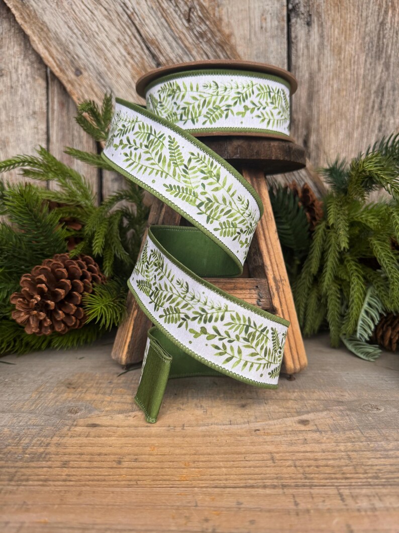 May include: A spool of wired ribbon featuring a white background with a repeating green watercolor fern leaf pattern. The ribbon has solid green finished edges and is coiled in a spiral shape, showcasing the botanical print on the front side.