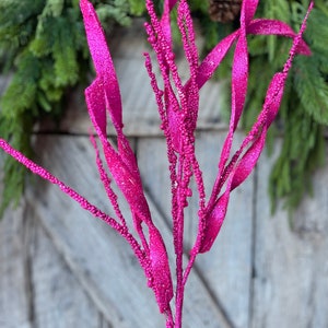 May include: A vibrant fuchsia decorative stem with glittery leaves and beaded accents. The artificial plant is set against a rustic wooden background and evergreen foliage, perfect for holiday or seasonal decor.