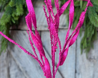May include: A vibrant fuchsia decorative stem with glittery leaves and beaded accents. The artificial plant is set against a rustic wooden background and evergreen foliage, perfect for holiday or seasonal decor.