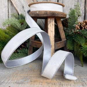 Puede incluir: Cinta de terciopelo blanco con borde plateado met&aacute;lico, presentada de forma decorativa. Una bobina de la cinta est&aacute; sobre un peque&ntilde;o taburete de madera. La cinta mide aproximadamente 2,5 cm de ancho.