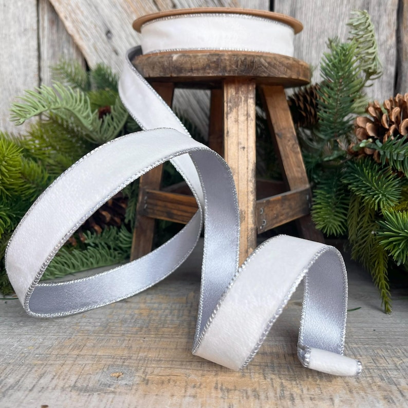 Puede incluir: Cinta de terciopelo blanco con borde plateado met&aacute;lico, presentada de forma decorativa. Una bobina de la cinta est&aacute; sobre un peque&ntilde;o taburete de madera. La cinta mide aproximadamente 2,5 cm de ancho.