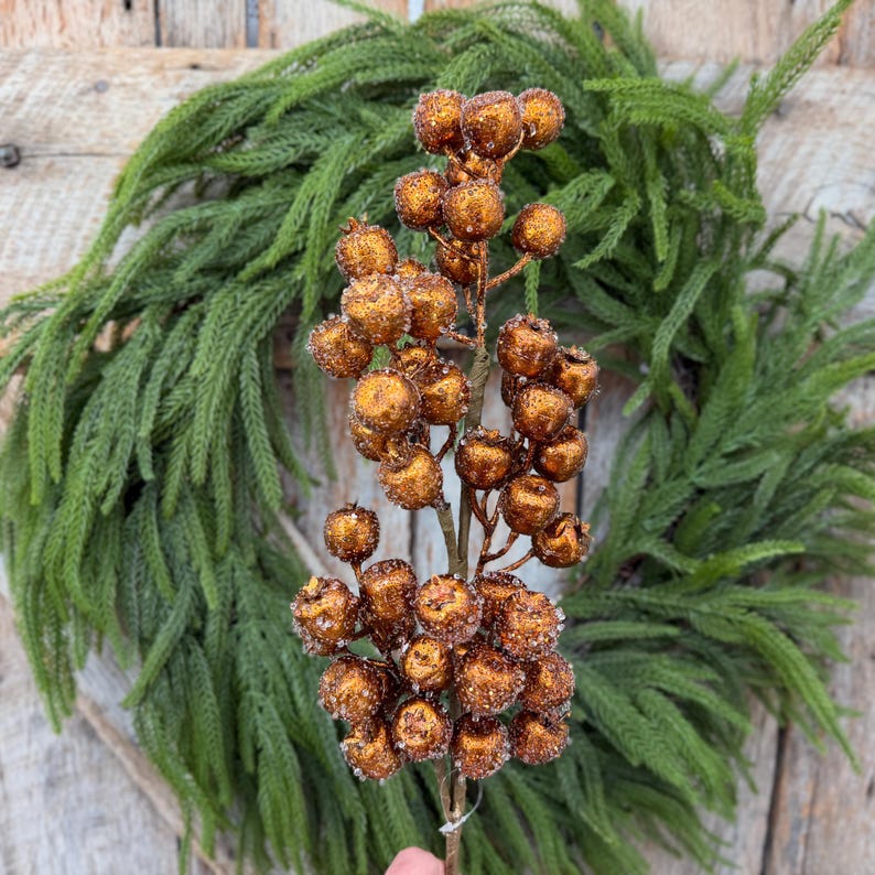 May include: A decorative floral pick featuring numerous round, metallic copper-colored berries with a textured, glittery finish. The berries are clustered along a central brown stem, creating a dense, branch-like structure with a shimmering appearance.