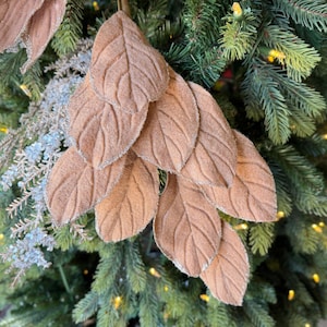 May include: A decorative spray of artificial leaves in a soft tan color. Each leaf features a textured, felt-like surface with visible veins and edges accented by a fine dusting of silver glitter, creating a subtle shimmer on the foliage.