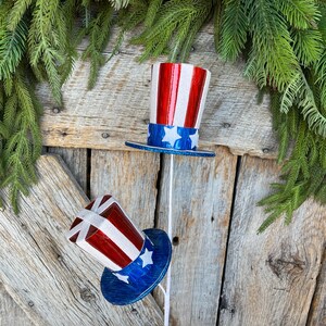 May include: Two red, white, and blue patriotic top hats with glitter on a wooden background. The hats are on white sticks.