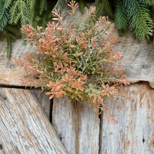May include: A close-up of a decorative floral arrangement featuring a cluster of orange and green faux plants. The arrangement is set against a rustic wooden background, with evergreen branches and pinecones in the background.