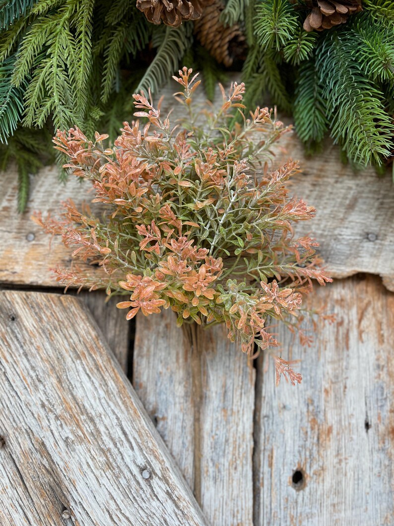 May include: A close-up of a decorative floral arrangement featuring a cluster of orange and green faux plants. The arrangement is set against a rustic wooden background, with evergreen branches and pinecones in the background.