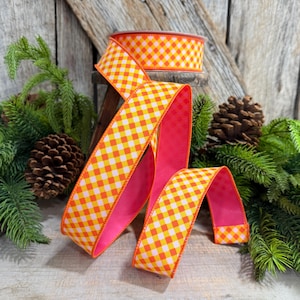 May include: A spool of wired ribbon featuring a vibrant orange and yellow gingham check pattern on the front side and a solid hot pink color on the reverse side. The ribbon is draped in loose folds, showcasing both the checkered and pink sides.