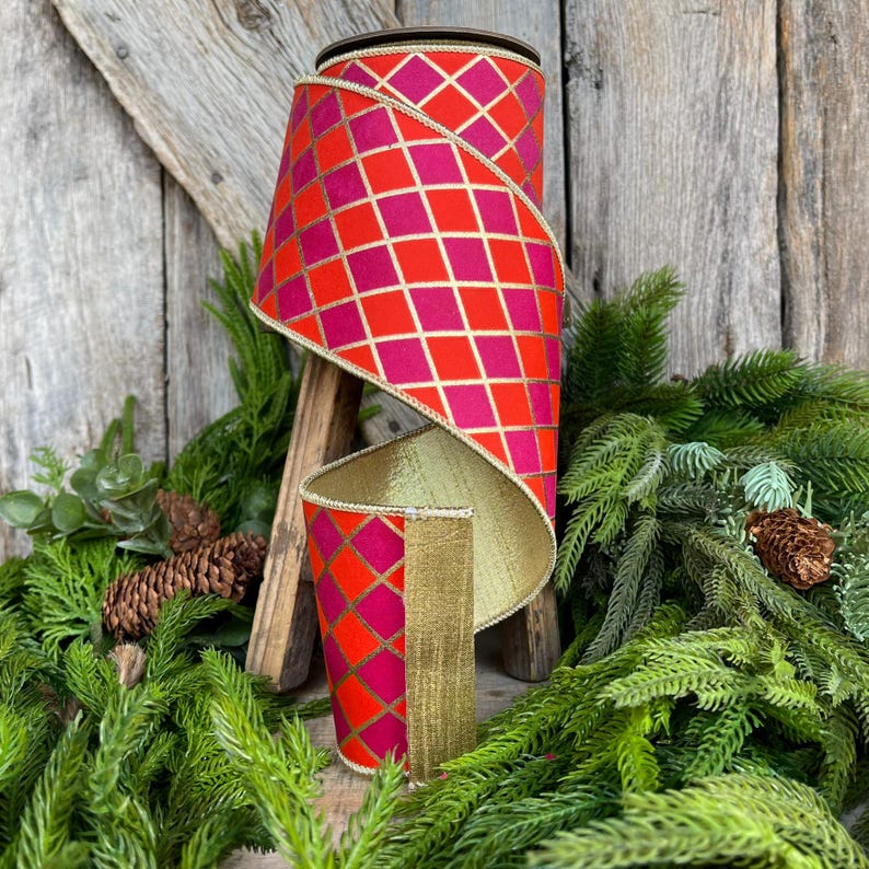 May include: A roll of decorative ribbon with a red and fuchsia diamond pattern, outlined in gold. The ribbon is displayed on a wooden stand, with a gold interior and gold trim. The background includes greenery and a wooden surface.