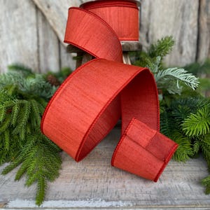 May include: A roll of wide, red ribbon with a textured appearance and a stitched edge. The ribbon is displayed with a flowing length, showcasing its color and texture. The ribbon is likely for gift wrapping or holiday decorations.