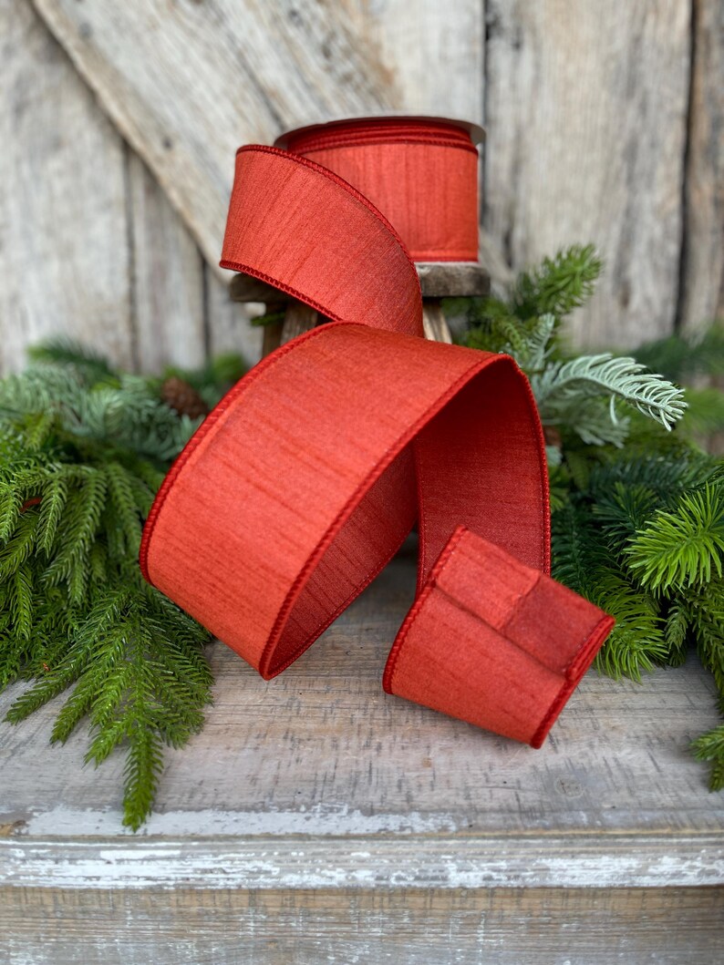 May include: A roll of wide, red ribbon with a textured appearance and a stitched edge. The ribbon is displayed with a flowing length, showcasing its color and texture. The ribbon is likely for gift wrapping or holiday decorations.