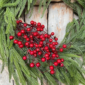 May include: A decorative wreath featuring lush green pine needles and a central cluster of glossy, vibrant red berries attached to thin, dark brown stems. The arrangement is circular, showcasing a dense texture of foliage and bright berry accents.