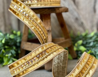 Op de afbeelding: Een rol gouden lint met een fluwelen textuur en een decoratieve rand van vierkante, amberkleurige strass-steentjes. Het lint wordt gepresenteerd op een houten krukje, met groen op de achtergrond.