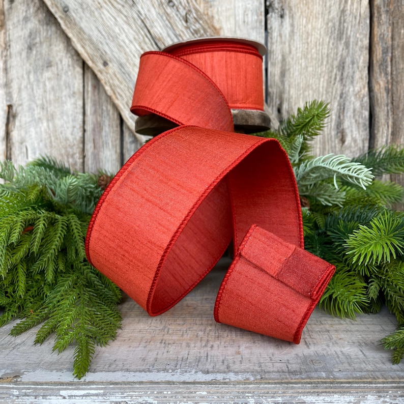 May include: A spool of red ribbon with a textured finish and a red border. A length of the ribbon is unrolled, and a small square of the ribbon is folded. The ribbon is displayed on a wooden surface with evergreen branches.