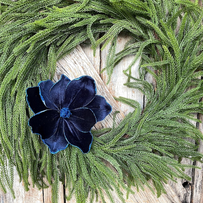 May include: A decorative wreath featuring a vibrant green foliage base with a large, navy blue velvet flower. The flower has a detailed design with a contrasting blue trim, adding a touch of elegance to the natural aesthetic. The wreath is set against a rustic wooden background.