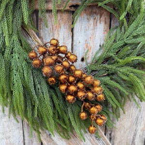 May include: A decorative wreath constructed from vibrant green evergreen foliage, featuring a central cluster of round, metallic bronze-colored berries with a textured, glittery finish that catches the light across the surface of the fruit.