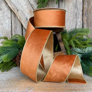 May include: A spool of wide, burnt orange velvet ribbon with a gold trim. The ribbon is partially unrolled, revealing a gold underside. The ribbon is displayed on a wooden surface with evergreen branches and a pinecone in the background.