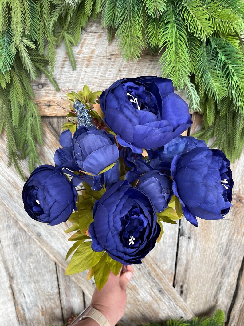 May include: A bouquet of deep blue artificial peonies with green leaves. The flowers are clustered together, held by a hand, against a rustic wooden background. Greenery frames the top of the image.