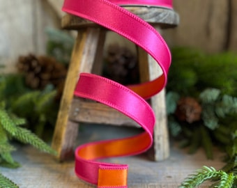 May include: A roll of bright pink satin ribbon with an orange underside, displayed on a small wooden stool. The ribbon is spiraled, showcasing its contrasting colors. The background includes greenery and a rustic wooden surface.