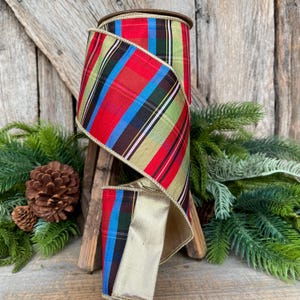 May include: A spool of decorative ribbon with a plaid pattern in red, green, blue, and black. The ribbon has a gold metallic backing and trim. The ribbon is displayed with pine branches and a pine cone, suggesting a holiday theme.