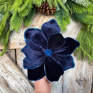 May include: A large, navy blue velvet flower with glittered edges. The flower is a decorative accent, possibly for holiday or seasonal decor. The background includes evergreen branches and a pinecone, suggesting a festive theme.