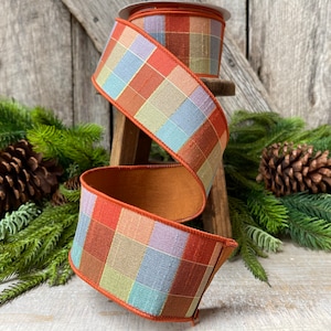 May include: A roll of decorative ribbon with a plaid pattern in shades of orange, red, green, blue, and purple. The ribbon has a brown border and is displayed on a small wooden stool. The background includes greenery and a pinecone.