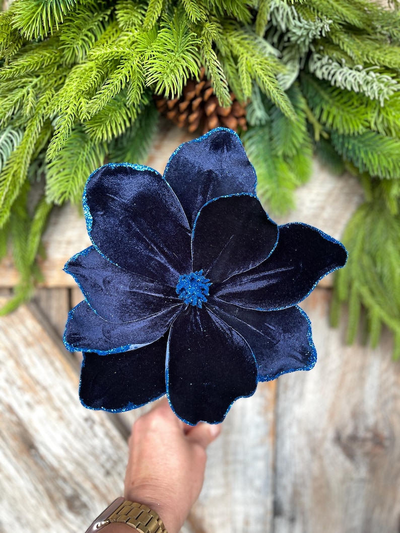 May include: A large, navy blue velvet flower with glitter edging and a textured center. The flower is held in front of a background of green pine needles and a pine cone. The flower's petals are layered, creating a full, dimensional look.