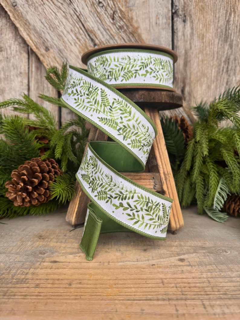 May include: A spool of wired ribbon featuring a white background with a repeating green fern leaf pattern. The ribbon has a solid green underside and finished edges. The design displays various shades of green foliage in a botanical style.