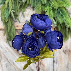 May include: A bouquet of deep blue artificial peonies with green leaves and stems. The flowers are in full bloom, with visible white centers. The background includes a green wreath and a wooden door.