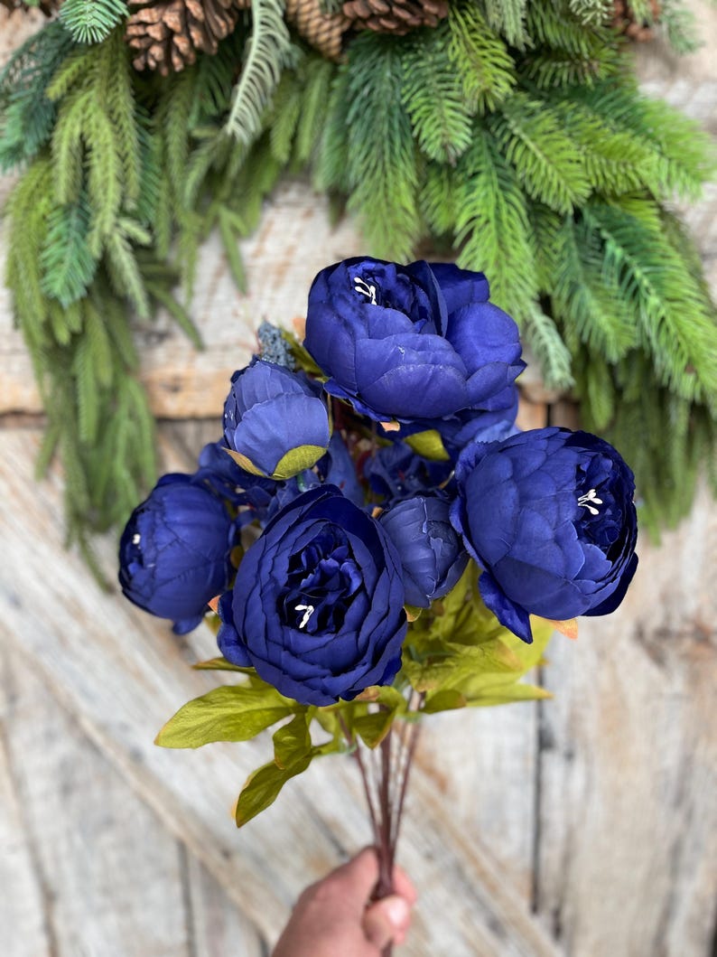 May include: A bouquet of deep blue artificial peonies with green leaves and stems. The flowers are in full bloom, with visible white centers. The background includes a green wreath and a wooden door.